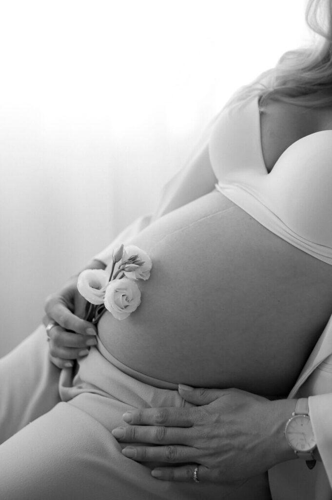 Schwarz-weiß Foto: Eine Frau hält eine weiße Rose vor ihren Babybauch beim Fotoshooting