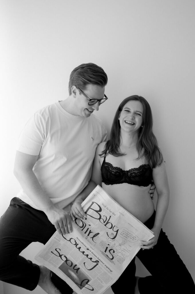 Eine schwangere Frau mit ihrem Mann beim Babybauchshooting mit einer Zeitung auf der steht "Babygirl is coming soon"